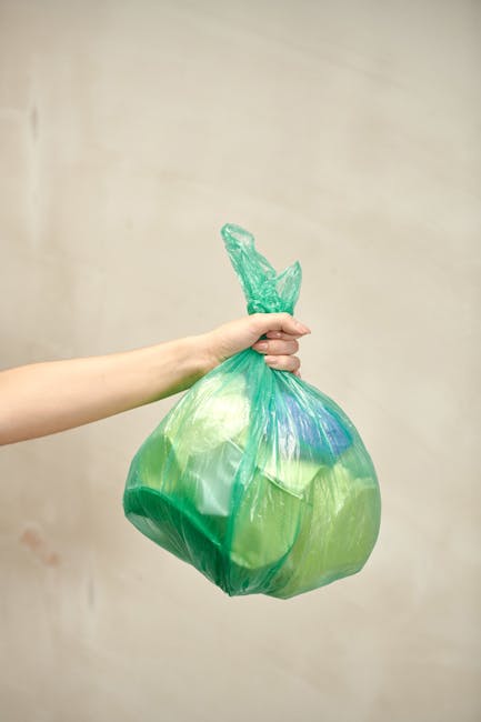 A person's hand and forearm are visible, holding a green plastic bag filled with rubbish, which appears to contain various waste materials. The bag is tightly knotted at the top, with some loose plast