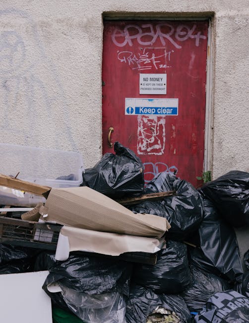 A person's hand and forearm are visible, holding a green plastic bag filled with rubbish, which appears to contain various waste materials. The bag is tightly knotted at the top, with some loose plast