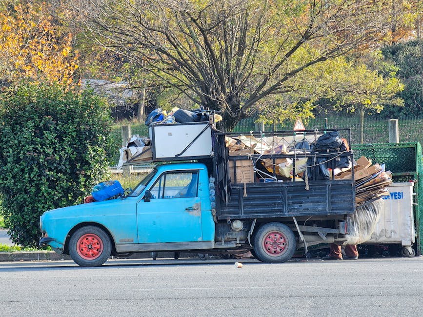 An outdoor scene showing a large pile of mixed household waste and recyclables on an urban sidewalk in front of a commercial building with blue metal railings and a tree on the left side. The waste in
