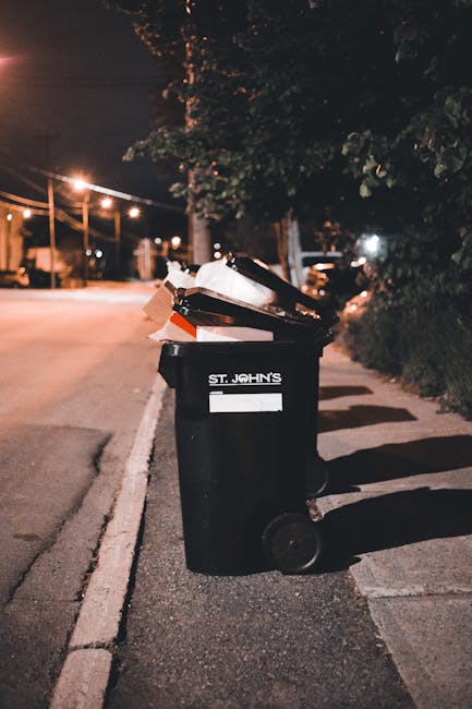 A black wheeled rubbish bin with the label 'St. John's' positioned on the edge of a paved residential street during nighttime. The bin is filled with various discarded items, including cardboard boxes